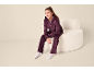 Fille en ensemble velours violet foncé avec sweat à capuche et pantalon, assise sur un fauteuil blanc.