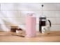 Moulin à café rose, cafetière à piston et grains de café sur un comptoir en bois.