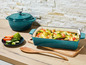 Un plat à gratin en fonte bleu avec des légumes cuits au four et une casserole en fonte plus petite.