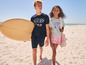 Deux enfants sur la plage, un garçon avec une planche de surf et une fille avec un sac.