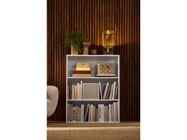 Une bibliothèque blanche à trois étagères remplie de livres et d'objets déco contre un mur en bois.