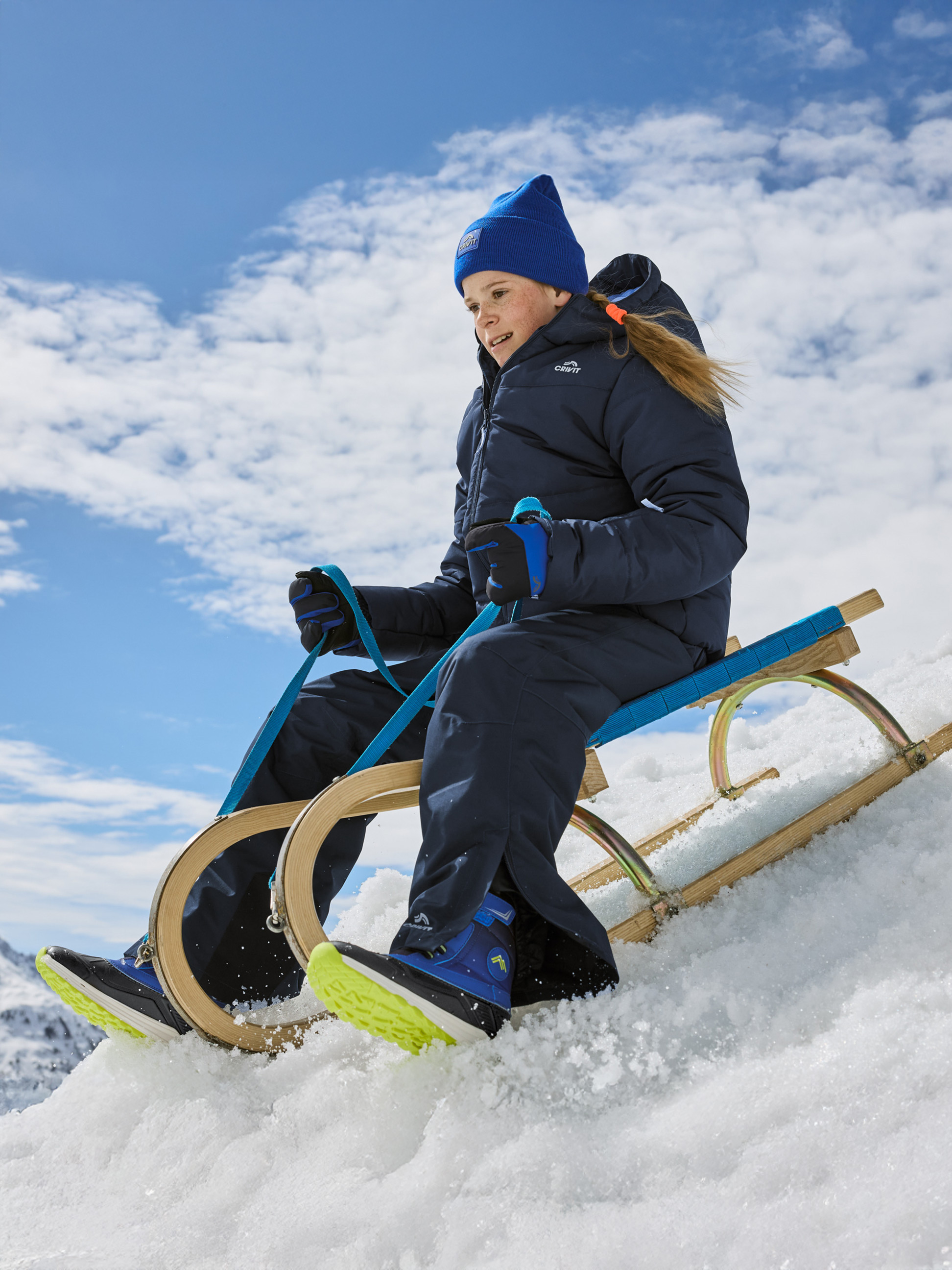 Fille en combinaison de ski et bonnet bleu faisant de la luge dans la neige.