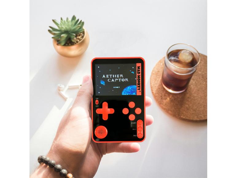 Main tenant une console de jeu noire et orange avec « AETHER CAPTOR », à côté d'une plante succulente et d'une boisson.