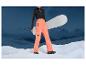 Une femme en pantalon de ski orange avec un snowboard et un paysage montagneux en arrière-plan.