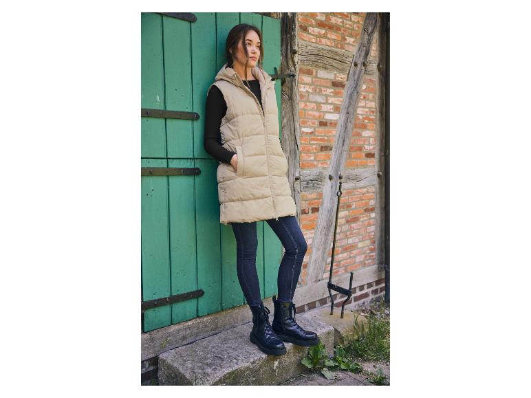 Femme en longue veste beige et bottes noires devant une porte en bois.