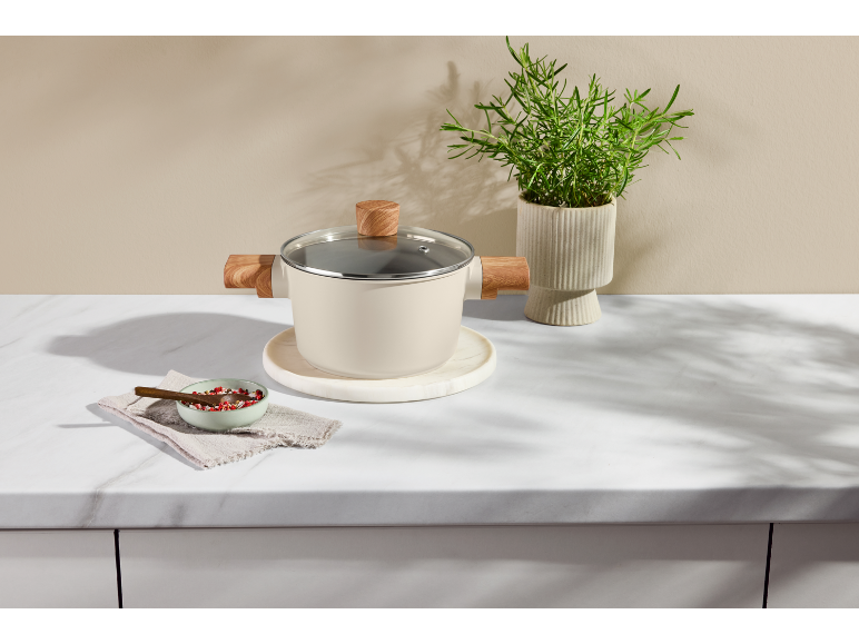 Casserole claire avec poignées en bois sur un plan de travail en marbre à côté d'une plante.