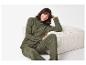 Femme souriante en pull et pantalon côtelés vert olive, assise à côté d'un pouf.