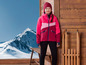 Une fille portant une veste de ski rose et un bonnet rouge se tient devant un chalet en bois.