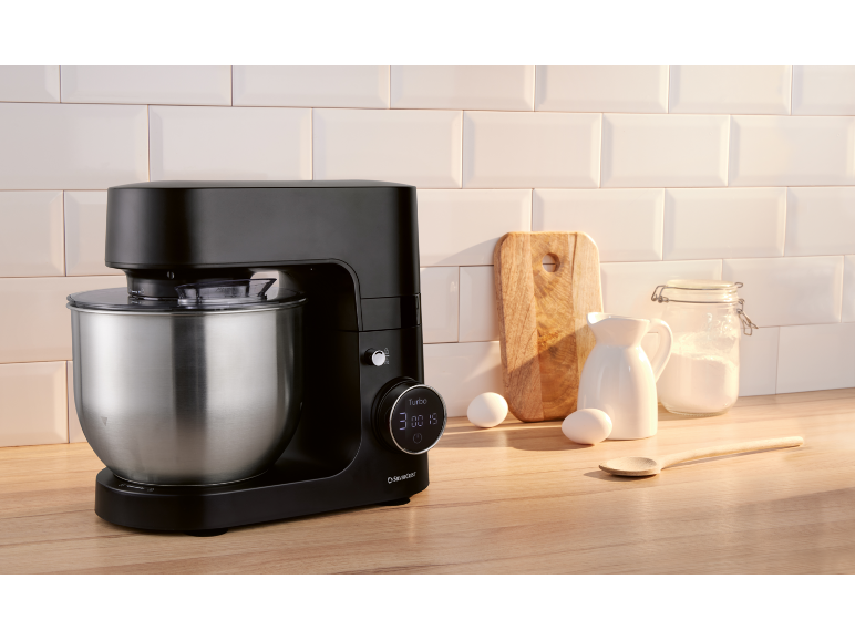 Robot pâtissier noir avec bol en acier inoxydable sur un comptoir en bois avec des ingrédients de cuisson.