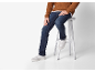 Homme en pull tricoté marron, jean bleu et baskets blanches, assis sur un tabouret.