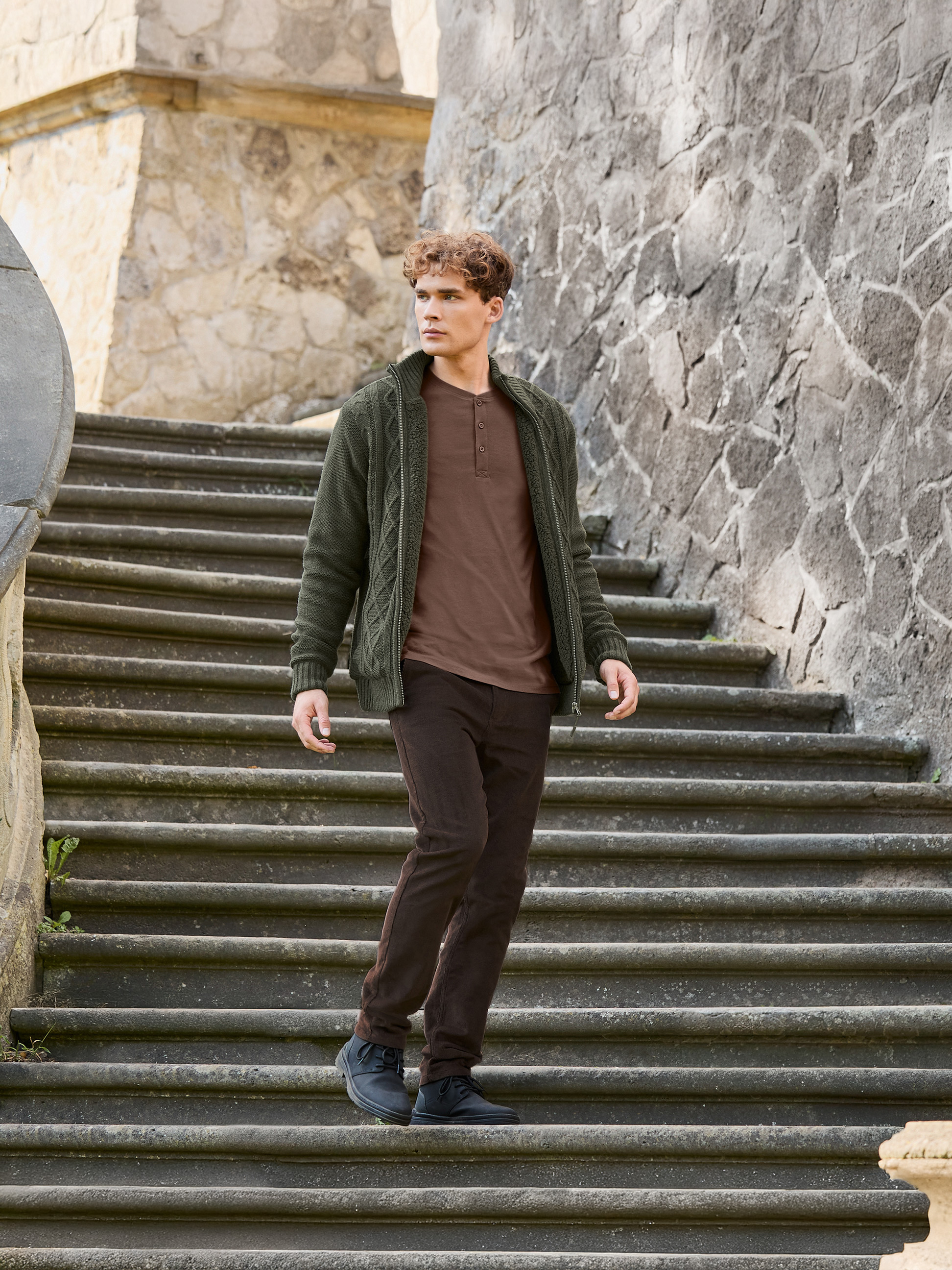 Homme en veste tricotée verte, T-shirt marron et pantalon marron descendant des escaliers en pierre.