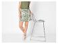Femme en débardeur vert et jupe à imprimé botanique, appuyée sur un tabouret de bar.