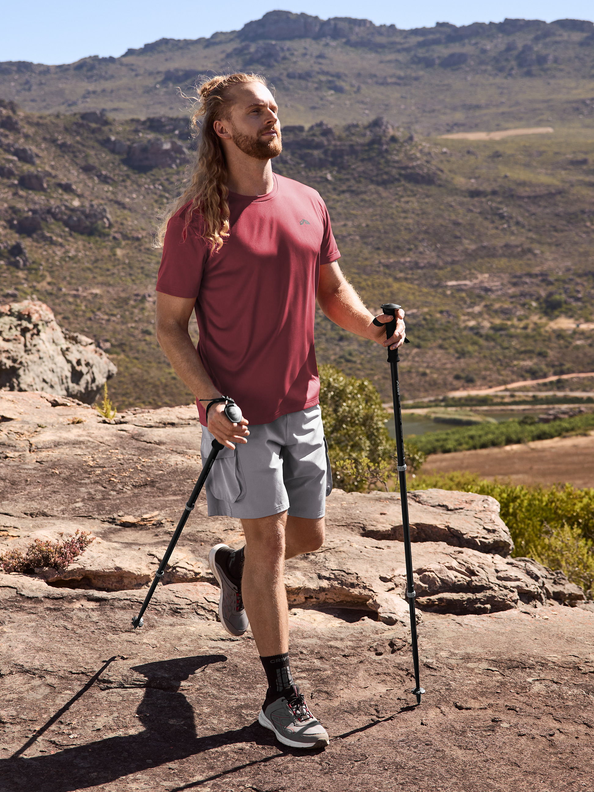 Homme en t-shirt et short de randonnée, avec bâtons de trekking dans un paysage montagneux.