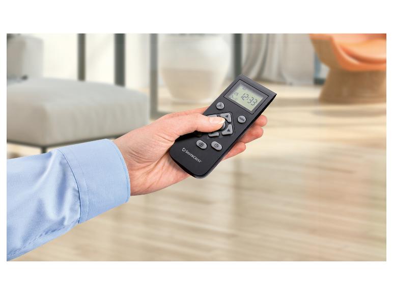 Une main tient une télécommande noire avec un écran numérique affichant 12:33, dans une pièce lumineuse.