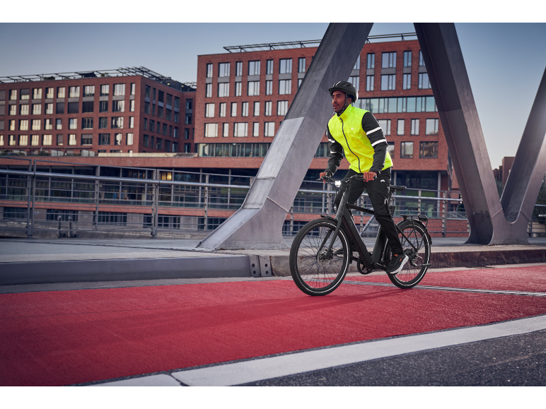 Homme en veste de cyclisme et casque roulant à vélo électrique sur une piste cyclable rouge.