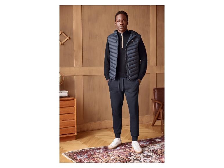 Homme en gilet bleu marine, pull et pantalon noirs, baskets blanches, sur un tapis à motifs.