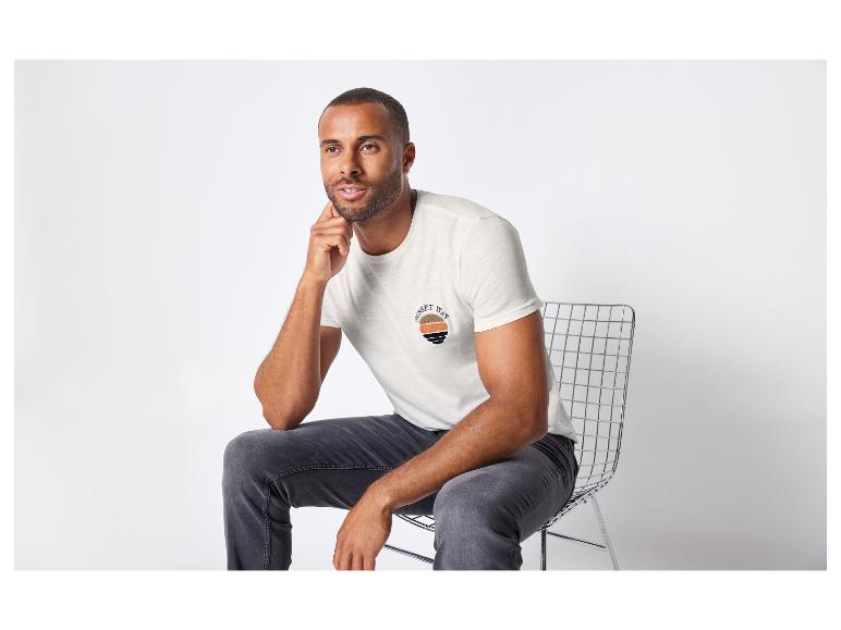 Homme avec un t-shirt blanc à imprimé coucher de soleil, porte un jean et est assis sur une chaise en métal.