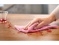 Une main nettoie un liquide rouge renversé sur une table en bois avec un chiffon rose.