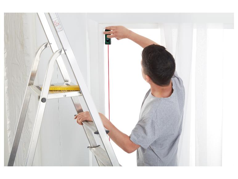 Un homme mesure une fenêtre avec un télémètre laser, debout sur une échelle.