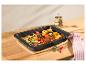 Plat de légumes rôtis avec pommes de terre, tomates et oignons dans un plat de cuisson noir sur une planche en bois.