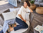 Une femme porte un t-shirt blanc avec un imprimé d'ancre et un pantalon noir, assise sur une chaise en rotin.