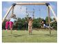 Enfants jouant sur une balançoire en bois avec anneaux et sièges, dans un jardin vert.