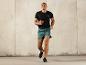 Un homme en t-shirt noir et short vert, courant en tenue de sport.
