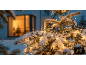 Lumières de Noël extérieures enneigées sur un arbre, avec une fenêtre chaleureusement éclairée en arrière-plan.