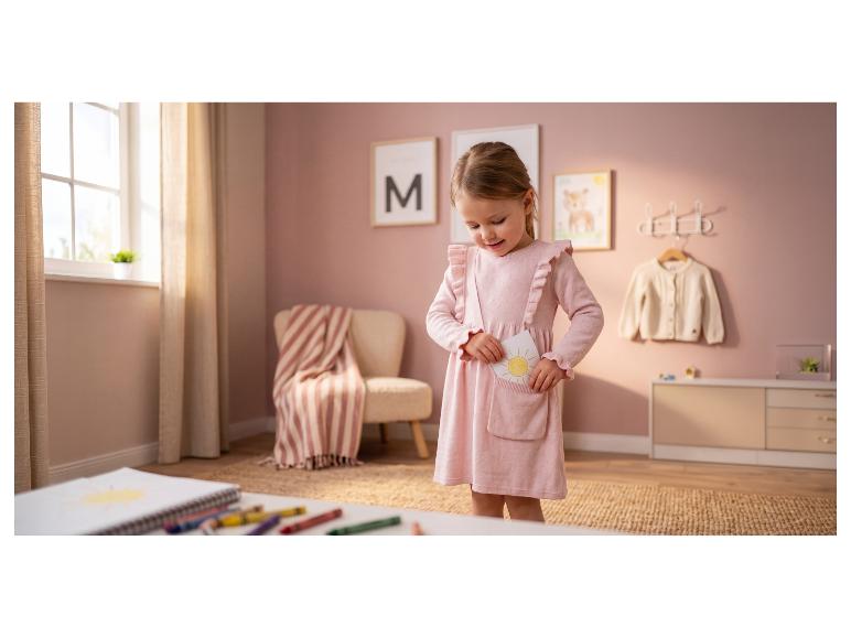 Fille en robe rose met un dessin de soleil dans sa poche, dans une chambre d'enfant.
