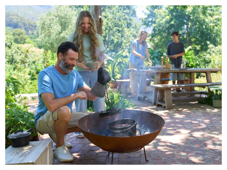 Famille cuisinant en extérieur avec une cocotte en fonte dans un brasero et une caisse en bois.