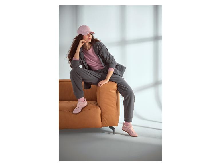 Femme en survêtement gris et casquette rose, assise sur un canapé marron.