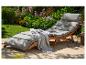 Chaise longue de jardin en bois avec coussins gris et couverture sur une terrasse fleurie.