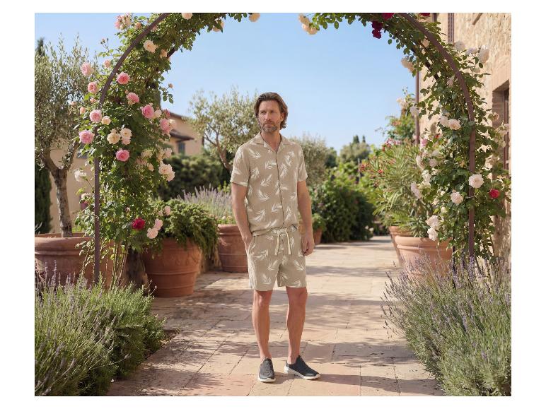 Homme en ensemble chemise et short beige à imprimé feuilles, dans un jardin.