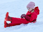 Une fille en combinaison de neige rouge avec un bonnet et des mitaines en fourrure, assise sur une luge dans la neige.