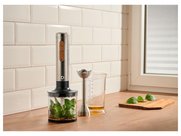 Mixeur plongeant avec basilic dans un récipient et verre doseur d'huile sur un comptoir en bois.
