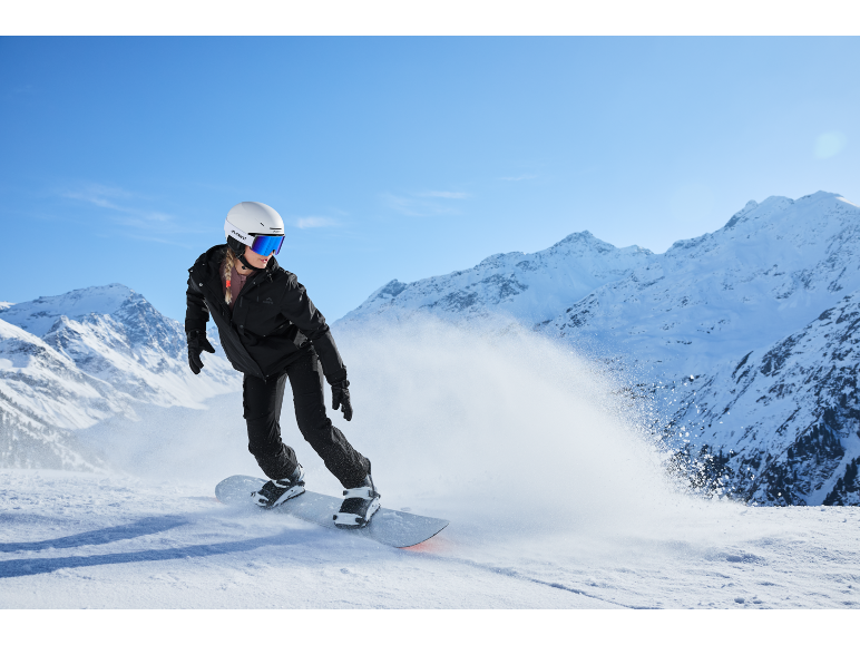 Une snowboardeuse en veste et pantalon de ski noirs, casque blanc et lunettes de ski bleues dévale une montagne enneigée.