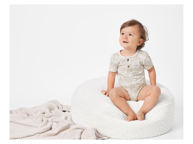 Bébé assis sur un pouf en fausse fourrure, vêtu d'un body à motifs tropicaux.