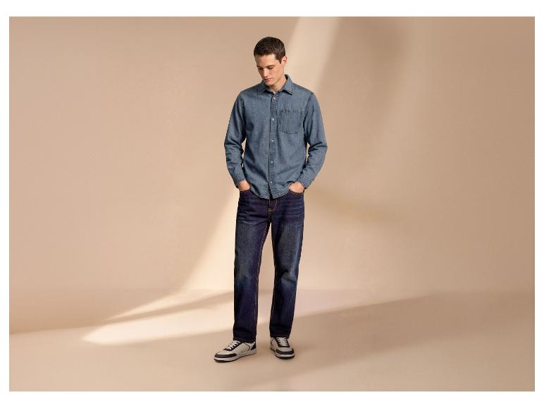 Homme en chemise et jean en denim, les mains dans les poches, regardant vers le bas.