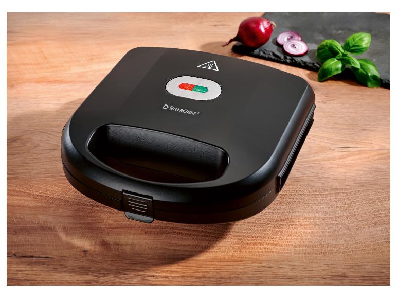 Appareil à croque-monsieur noir SilverCrest sur une table en bois avec oignon et basilic.