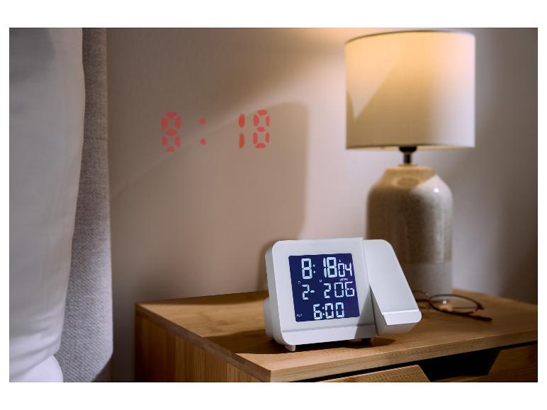 Réveil à projection blanc sur une table de chevet en bois avec une lampe.