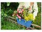 Personne tenant une jardinière bleue avec des fleurs roses et jaunes, au-dessus d'une clôture en fer.