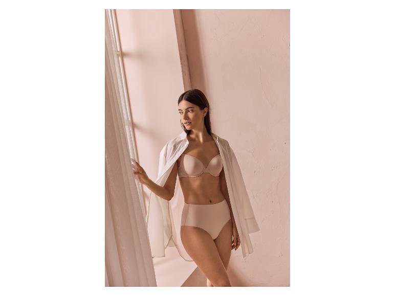 Femme en soutien-gorge et culotte beiges, avec une chemise blanche, près d'une fenêtre.