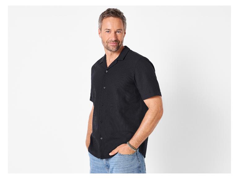 Homme en chemise noire à manches courtes et jean, posant les mains dans les poches.
