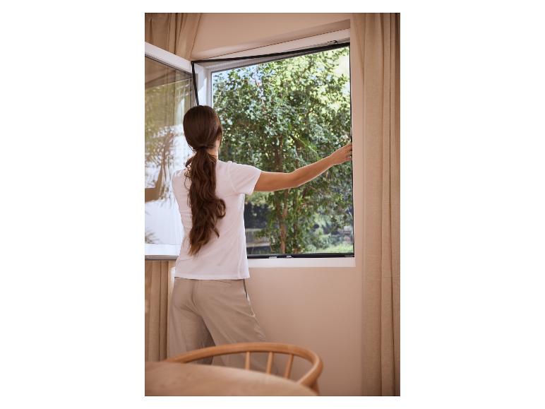 Femme ouvrant une fenêtre avec moustiquaire, verdure à l'extérieur.