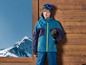 Un garçon porte une veste de ski bleue avec une fermeture éclair verte et un pantalon de ski bleu sur un fond de bois.