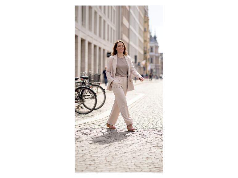 Femme en costume clair marchant dans une rue pavée, souriante.