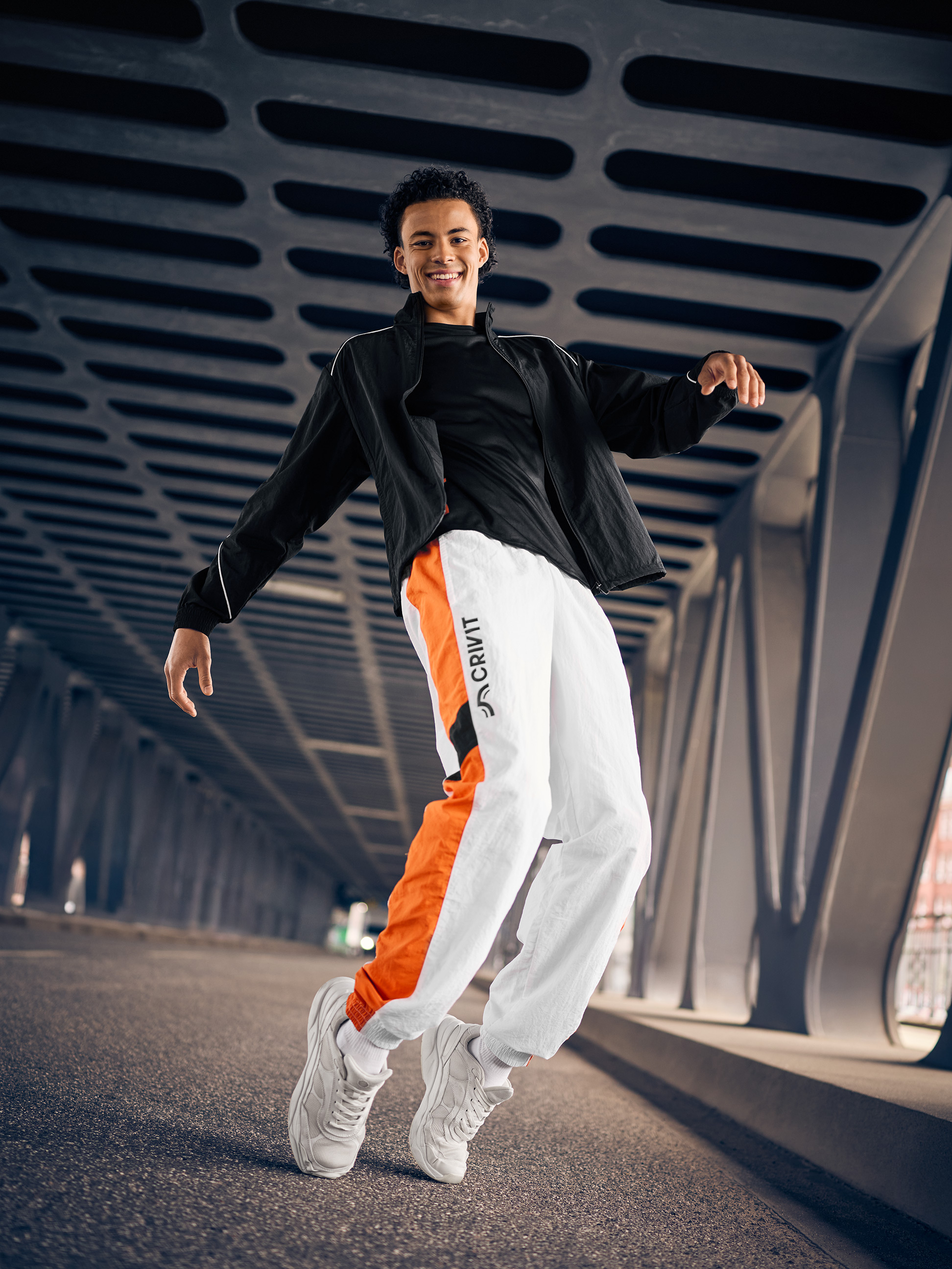 Homme en veste d'entraînement noire et pantalon d'entraînement blanc avec détails orange, sautant sur un pont.