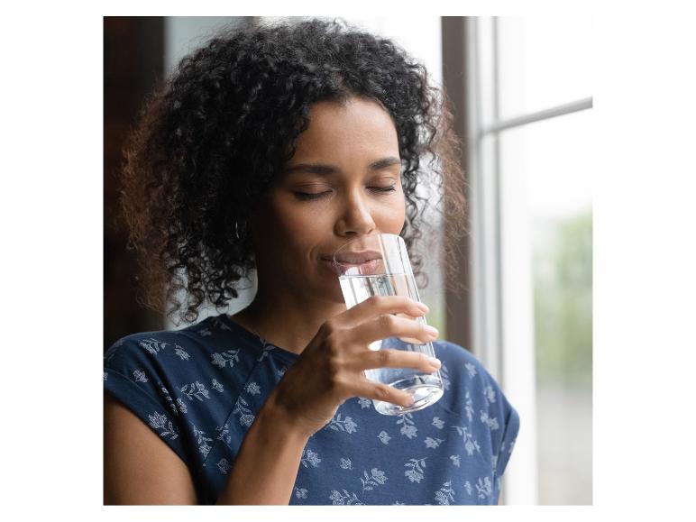 Femme aux cheveux bouclés buvant de l'eau dans un verre, vêtue d'un chemisier bleu fleuri.