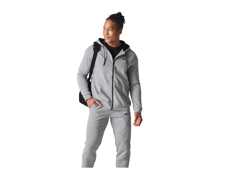 Homme en survêtement gris avec un t-shirt noir et un sac à bandoulière noir, regardant à droite.