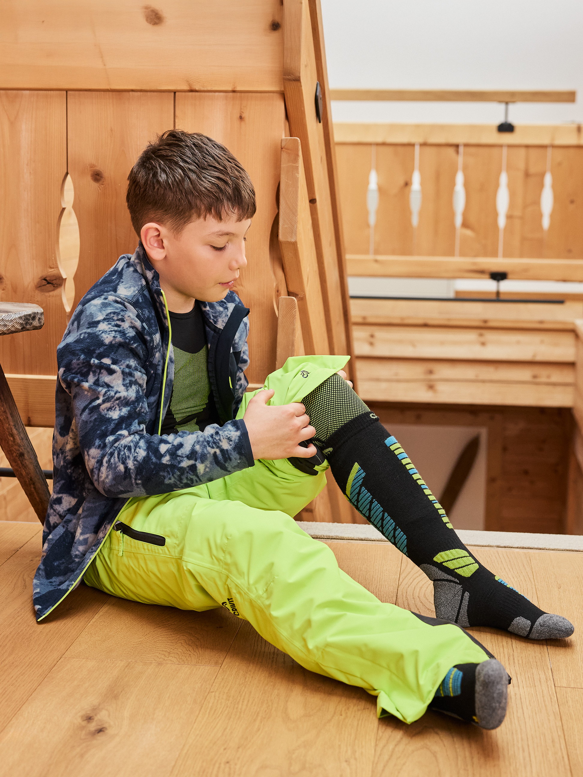 Enfant assis portant un pantalon de ski vert fluo et des chaussettes de ski noires.